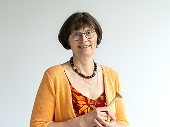 Frau mit kurzen braunen Haaren, Brille, oranger Strickjacke und gemustertem Kleid, die ein Holzobjekt in den Händen hält und lächelt