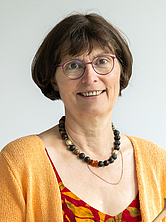 Frau mit kurzen braunen Haaren, Brille, oranger Strickjacke und gemustertem Kleid, die ein Holzobjekt in den Händen hält und lächelt