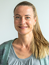 Frau mit langem, zurückgebundenem Haar lächelt in die Kamera und trägt einen grauen Pullover und eine grüne Strickjacke
