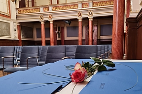 Blaue Zeugnismappen mit der Aufschrift 'URKUN' liegen auf einem Tisch in einem prunkvollen Saal, darauf liegt eine einzelne rote Rose