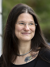 Frau mit langen braunen Haaren, Brille und Halsreif, die lächelnd nach rechts schaut