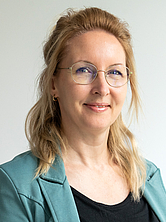 Frau mit langen blonden Haaren, Brille und grünem Blazer lächelt vor hellem Hintergrund