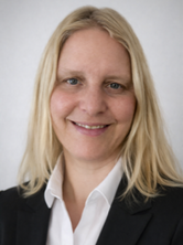 Porträt einer lächelnden Frau mit langen blonden Haaren, weißem Hemd und dunklem Blazer vor hellem Hintergrund