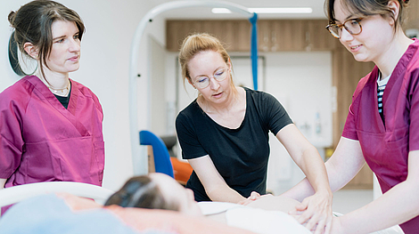 Drei Frauen, zwei in medizinischer Arbeitskleidung, üben an einer liegenden Person eine Behandlung oder Untersuchung in einem hellen Raum