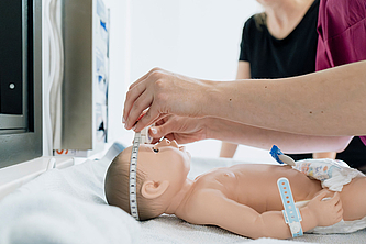 Hand misst mit einem flexiblen Maßband den Kopfumfang einer Babypuppe, die auf einer weißen Unterlage liegt