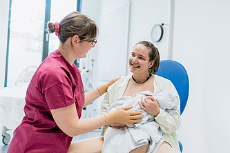 Frau im weißen Bademantel hält ein in eine Decke gewickeltes Baby und lächelt eine in roter Arbeitskleidung und Handschuhen gekleidete Frau an