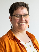 Lächelnde Frau mit kurzen dunklen Haaren, Brille und silbernen Ohrringen trägt ein orangefarbenes Hemd über einem weißen Shirt mit floralem Muster vor hellem Hintergrund