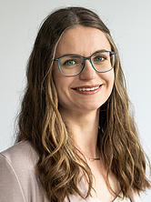Frau mit langen braunen Haaren, Brille und hellbeiger Jacke lächelt vor hellem Hintergrund